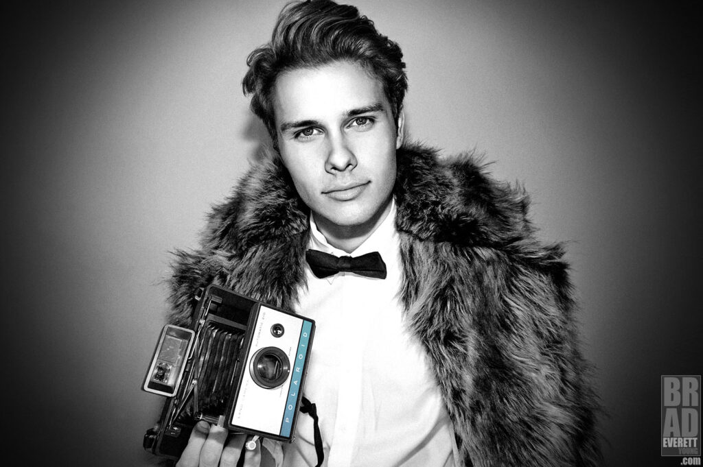 photo of Logan Shroyer in a faux fur coat holding a vintage polaroid camera